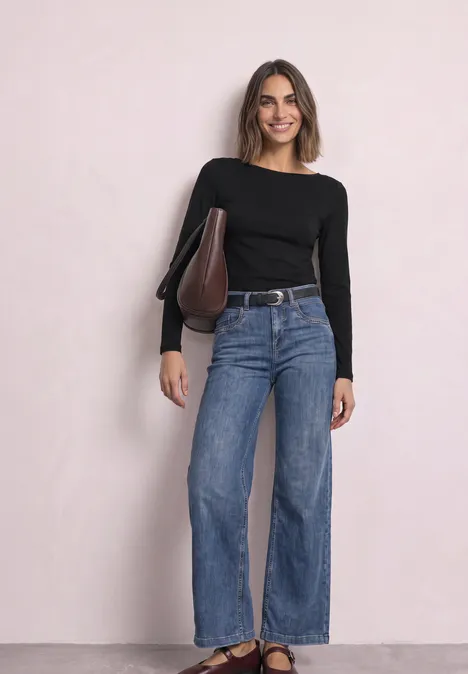 Jeune femme souriante devant un fond clair, pull noir, jean large et sac marron sur l’épaule.