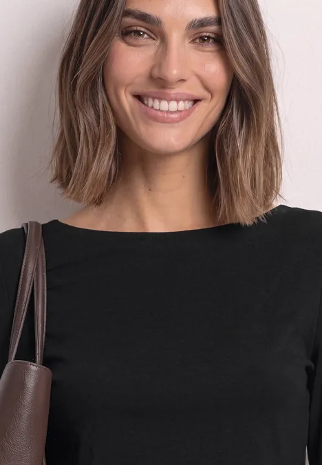 Jeune femme souriante en haut noir avec sac à bandoulière marron sur fond clair.
