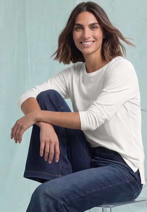 Langarm Shirt mit U-Boot-Ausschnitt off white Eine lächelnde Frau im weißen Langarmshirt und Jeans sitzt entspannt auf einem Hocker.
