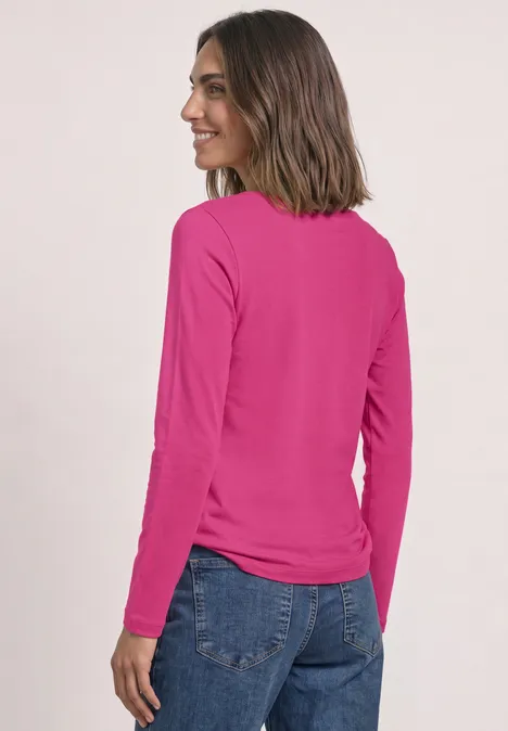 Een vrouw met halflang bruin haar draagt een roze longsleeve en een blauwe broek en glimlacht zijwaarts.