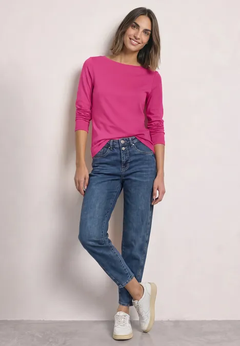 Vrouw leunt nonchalant tegen muur, roze trui, blauwe jeans en witte sneakers.