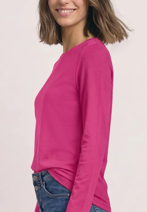 Vrouw in roze shirt met lange mouwen en spijkerbroek, zijwaarts poseerend, glimlachend voor lichte achtergrond.