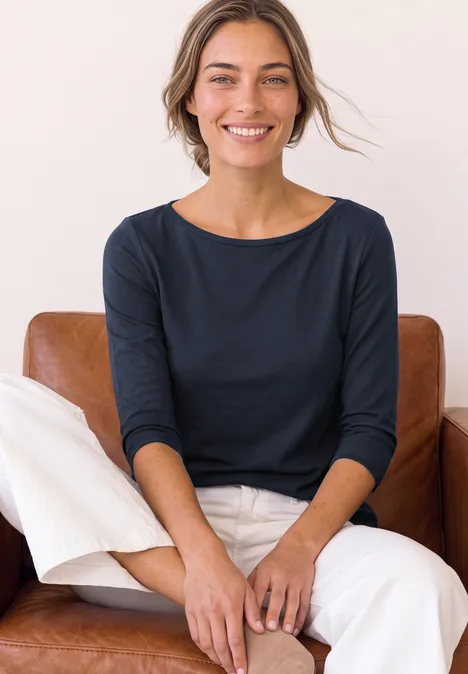 Langarm Shirt mit U-Boot-Ausschnitt darkest blue Eine Frau sitzt auf einem braunen Ledersofa, lächelt und trägt ein dunkelblaues Oberteil mit weißen Hosen.