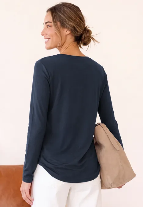 Langarm Shirt mit U-Boot-Ausschnitt darkest blue Frau in dunkelblauem Langarmshirt und weißer Hose hält beige Jacke und blickt seitlich, Rückansicht.