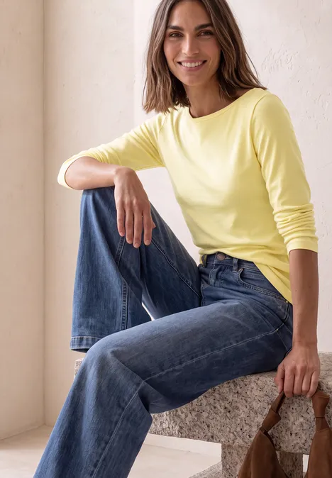 Frau sitzt lächelnd auf einem Block, trägt gelbes Langarmshirt, blaue Jeans und hält eine braune Tasche.