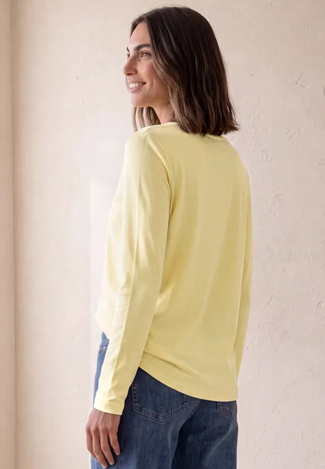 Frau in gelbem Langarmshirt, seitlich stehend, blickt nach links vor hellem Hintergrund.