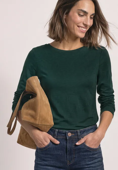 Frau lächelt, trägt dunkelgrünen Pullover, Jeans und hat eine braune Wildledertasche über der Schulter.