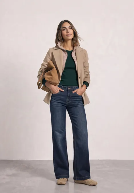 Eine Frau steht mit blauen Flared-Jeans, schwarzem Oberteil und beiger Jacke, hält eine Papiertüte.