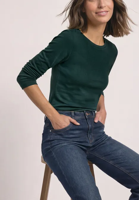 Frau sitzt auf einem Hocker, trägt grün langärmliges Shirt und blaue Jeans, Hände in den Taschen.