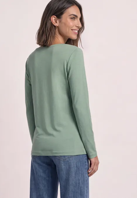 Une femme de profil porte un haut vert à manches longues et un jean bleu en souriant.