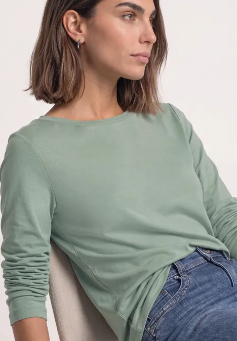 Femme photographiée de profil portant un t-shirt à manches longues vert menthe et un jean, assise.