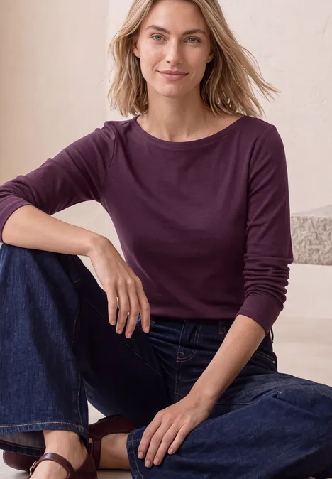 Langarm Shirt mit U-Boot-Ausschnitt berry maroon Frau sitzt entspannt in Jeans und langärmeligem burgunderfarbenem Shirt, lächelnd vor hellem Hintergrund.