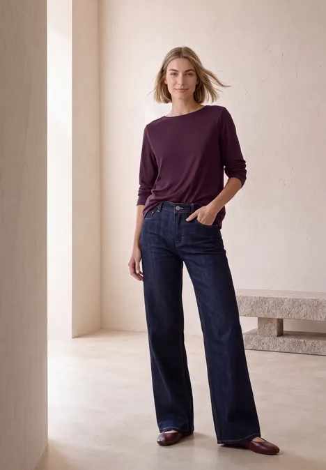 Langarm Shirt mit U-Boot-Ausschnitt berry maroon Frau in weiten Jeans und pflaumenfarbenem Longsleeve vor minimalistischem, hellem Hintergrund.