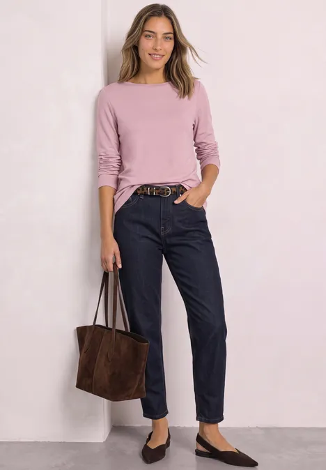 Femme en haut manches longues rose, jean noir, chaussures plates et sac marron devant un mur clair.