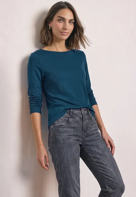 Frau in blau-grünem Langarmshirt und grauen Jeans steht lächelnd vor neutralem Hintergrund.