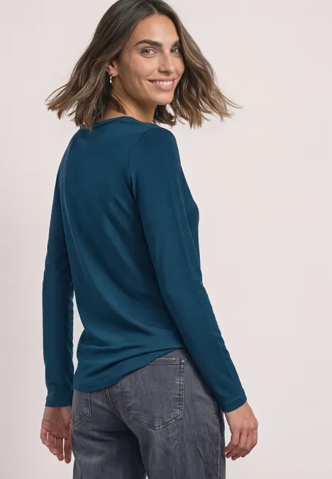 Frau in blauem Langarmshirt und dunkler Jeans lächelt und blickt seitlich über die Schulter.
