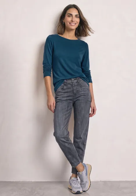 Eine Frau trägt ein blaugrünes Shirt, graue Jeans und weiße Sneakers vor hellem Hintergrund.