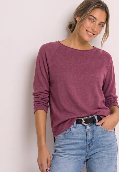 Junge Frau in violettem Pullover und Jeans, lächelt entspannt, eine Hand in der Hosentasche.