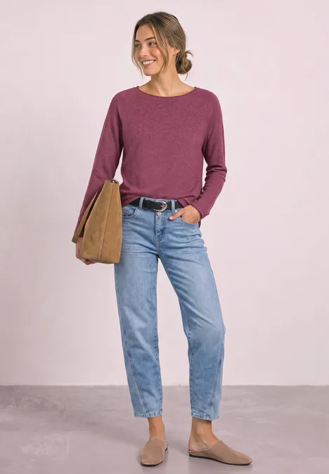 Frau in lila Pullover, Jeans und braunen Stiefeletten trägt Tasche und lächelt.