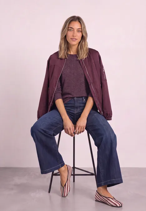 Femme assise sur un tabouret devant un mur rose pâle, vêtue d’une veste bordeaux, jean large et sandales rayées.