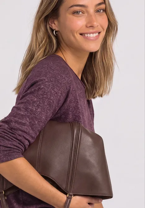 Une femme souriante en pull violet porte sur le côté un sac à main en cuir marron.