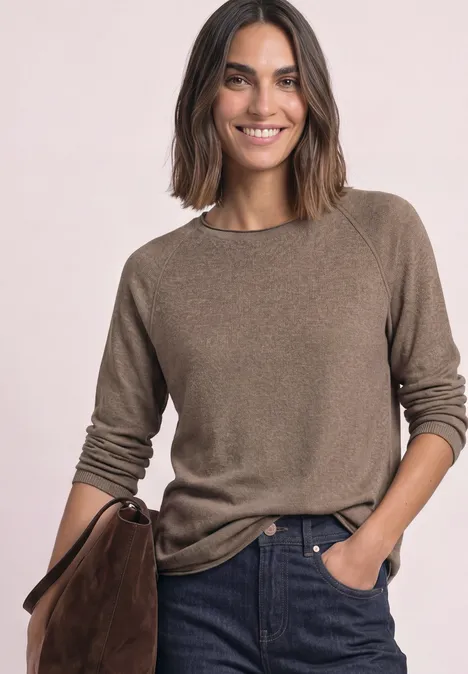 Femme aux cheveux mi-longs porte un pull marron, un jean et tient un sac marron tout en souriant.