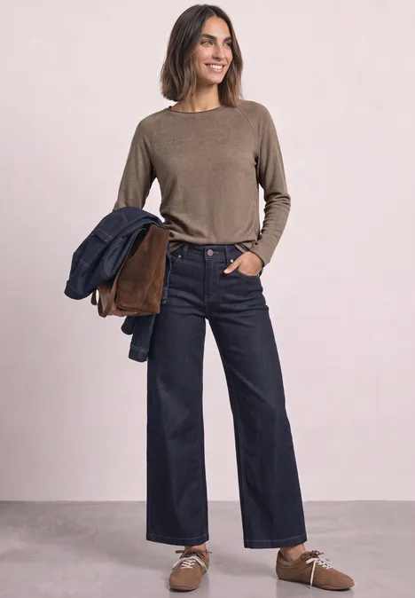 Femme en pull marron et jean large, tenant des vestes sur le bras.