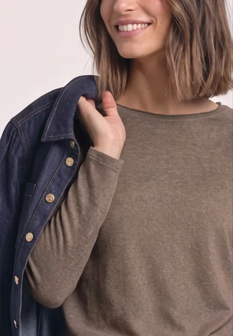 Femme en haut marron à manches longues, portant une veste en jean sur l’épaule, souriante.