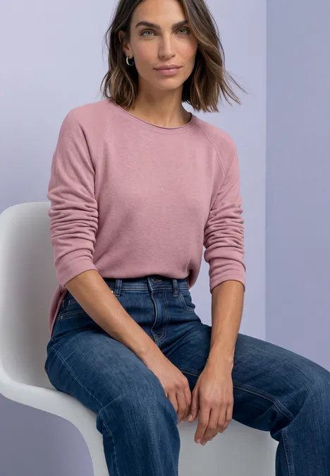 T-shirt à manches longues, col rond et bord roulé vintage blush mel. Eine Frau sitzt auf einem weißen Stuhl, trägt einen rosa Pullover und blaue Jeans.