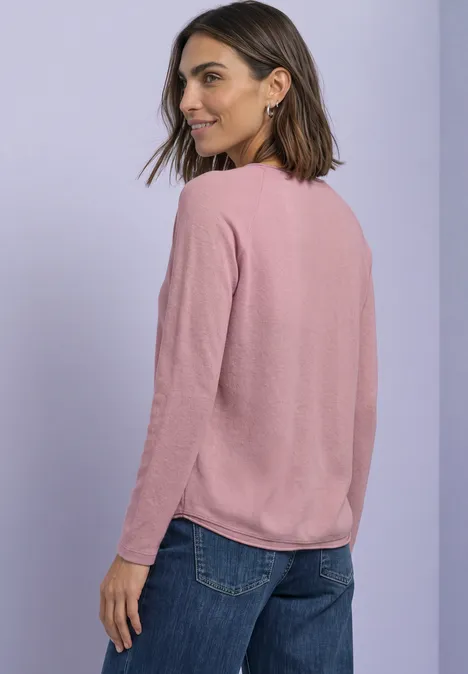 T-shirt à manches longues, col rond et bord roulé vintage blush mel. Frau mit rosa Pullover und blauer Jeans blickt über die Schulter vor hellem Hintergrund.