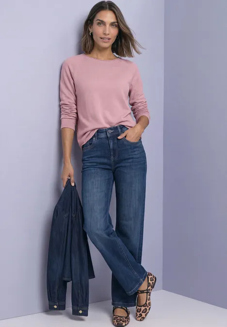 T-shirt à manches longues, col rond et bord roulé vintage blush mel. Frau im rosa Pullover, blauen Jeans und Leoparden-Sandalen hält eine Jeansjacke vor lila Hintergrund.