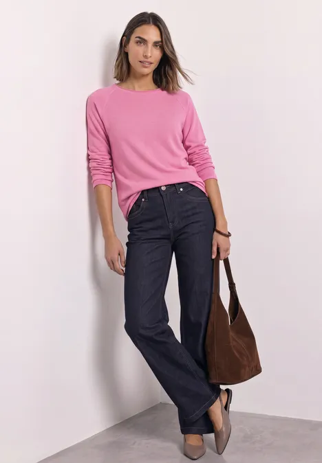 Eine Frau lehnt lässig an einer Ecke, trägt ein pinkes Oberteil, dunkelblaue Jeans und hält eine braune Tasche.