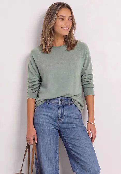 Eine lächelnde Frau in grünem Pullover und blauen Jeans hält eine Tasche.