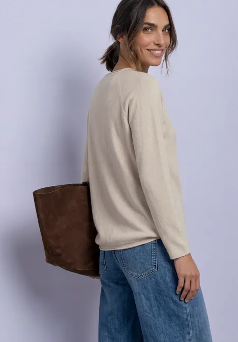 T-shirt à manches longues, col rond et bord roulé clay sand mel. Eine Frau mit beigem Pullover, Jeans und brauner Tasche blickt lächelnd über die Schulter.