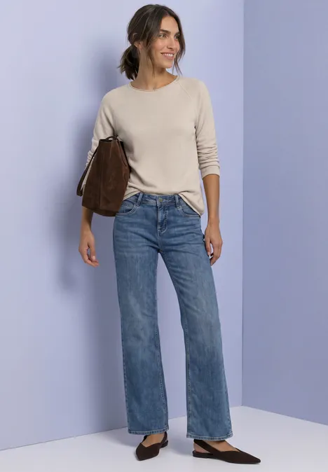 T-shirt à manches longues, col rond et bord roulé clay sand mel. Eine Frau in beigem Pullover und weiten Jeans mit brauner Tasche vor pastellfarbenem Hintergrund.