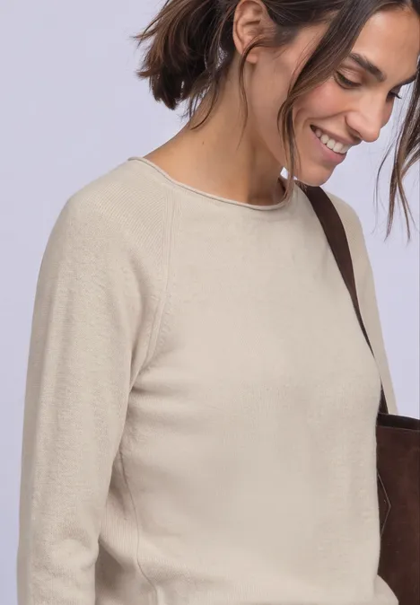 T-shirt à manches longues, col rond et bord roulé clay sand mel. Fröhliche Frau in beigem Pullover mit dunkelbraunem Schulterriemen vor neutralem Hintergrund.