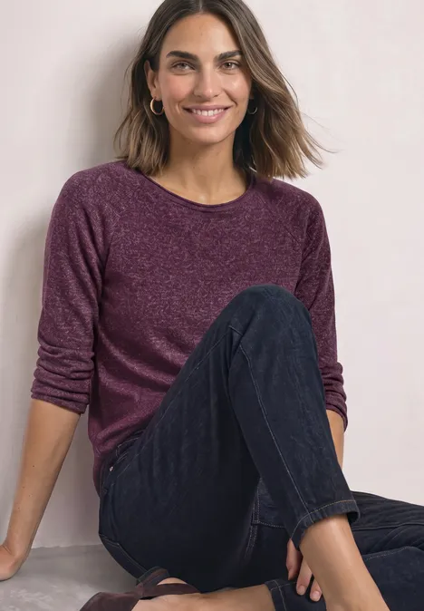Eine lächelnde Frau sitzt entspannt und trägt einen violetten Pullover mit dunklen Hosen.