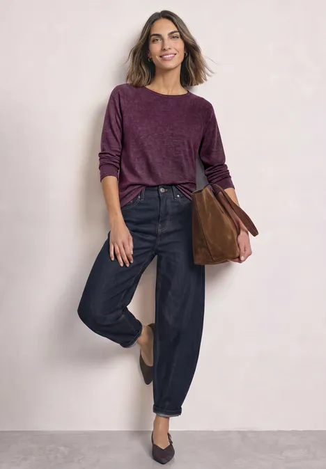 Eine lächelnde Frau trägt einen weinroten Pullover, schwarze Jeans und eine braune Tasche.