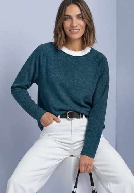 Frau sitzt vor hellem Hintergrund, trägt blaugrünen Pullover, weiße Hose und lächelt.
