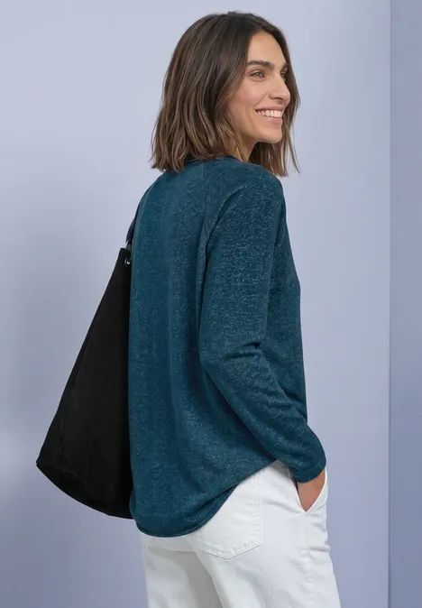 Frau in grünem Pullover und weißer Hose, lächelnd über die Schulter blickend, mit einer schwarzen Tasche.