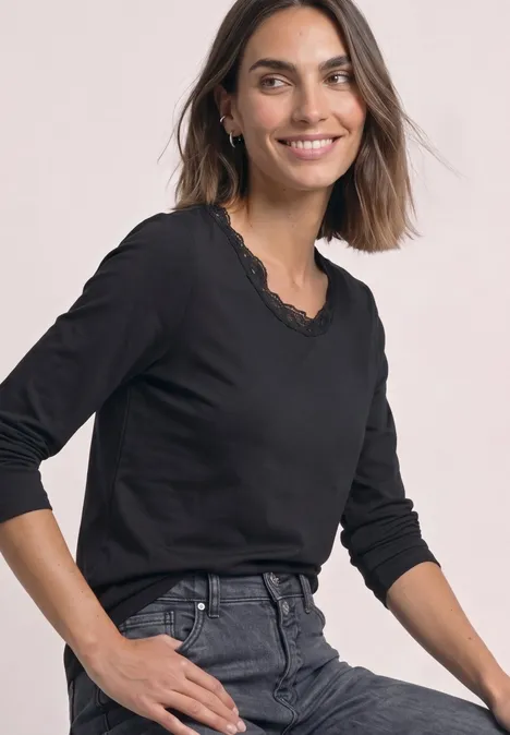 Une femme souriante en haut noir à manches longues et détail en dentelle, assise devant un fond clair.