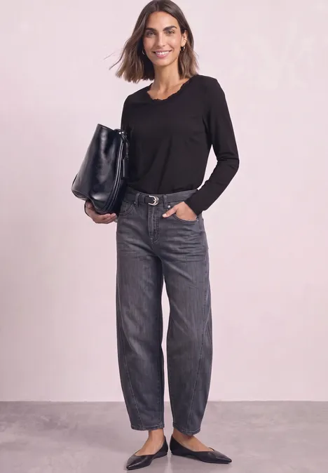 Femme en haut noir à manches longues, jean ample gris et ballerines noires, tenant un grand sac noir devant un mur clair