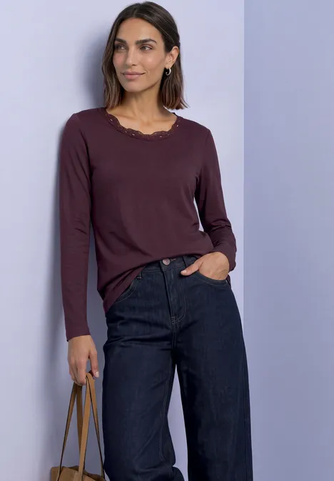 Frau in weinrotem Langarmshirt und dunklen, weiten Jeans hält eine Handtasche.