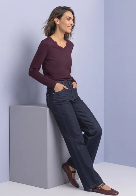 Frau lehnt an grauem Podest, trägt weinrotes Langarmshirt, weite dunkle Jeans und braune Schuhe vor hellem Hintergrund.