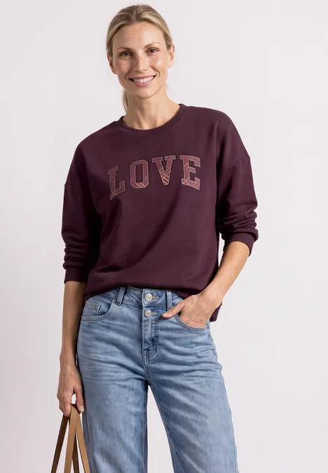Femme souriante en sweat bordeaux «LOVE», jean taille haute, tenant un sac à main.