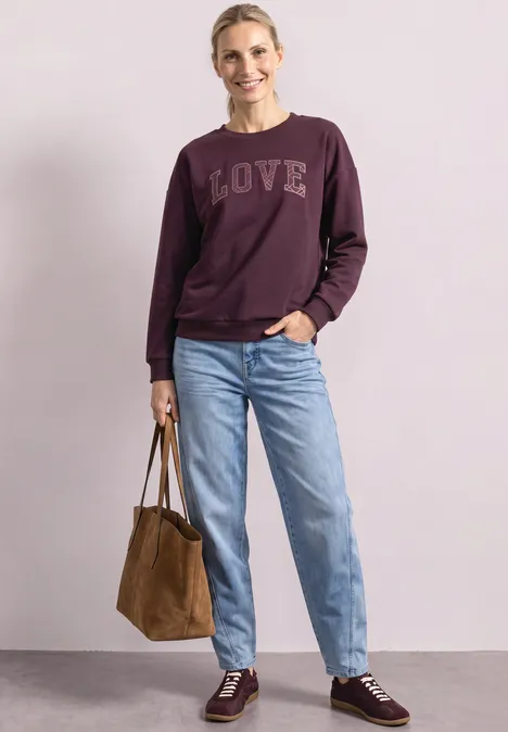 Femme souriante devant un fond clair, sweat bordeaux LOVE, jean bleu, baskets assorties et sac marron.
