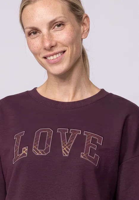 Femme blonde souriante aux cheveux attachés en queue de cheval, vêtue d'un sweat violet avec l'inscription 'LOVE'.