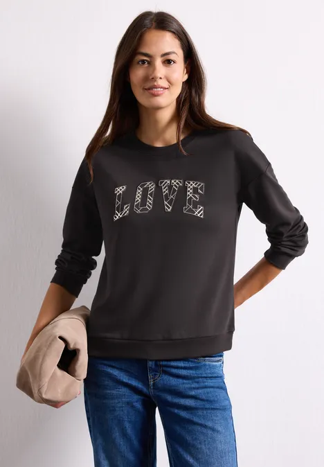 T-shirt à manches longues avec motif typographique black coffee Frau trägt schwarzen Pullover mit LOVE-Schriftzug, kombiniert mit blauen Jeans und hält eine beige Jacke.