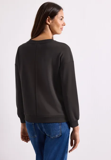 T-shirt à manches longues avec motif typographique black coffee Frau steht mit dem Rücken zur Kamera, trägt schwarzen Pullover und blaue Jeans vor weißem Hintergrund.