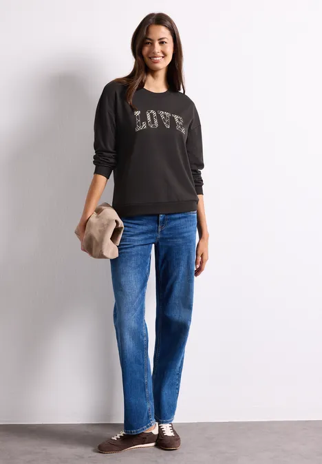 T-shirt à manches longues avec motif typographique black coffee Junge Frau im schwarzen Pullover mit LOVE-Schriftzug und blauen Jeans, lächelt und hält einen beigen Hut.